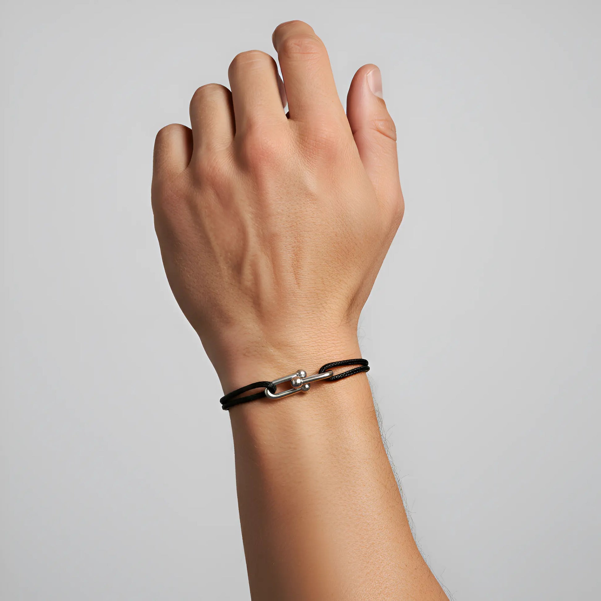 Hand wearing a black leather bracelet with a metal clasp on a light gray background